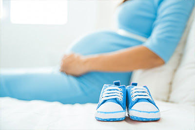 Blue baby sneakers with white laces on a white couch next to a pregnant woman wearing a blue dress.
